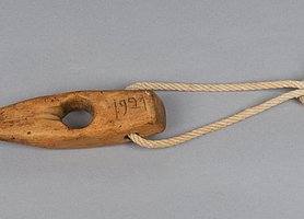 Das Seilholz sieht aus wie ein Keil. In der Mitte ist ein Loch. An dem Loch sind Spuren von Seilen. Auf der Seite steht die Jahreszahl 1921. In einem weiteren Loch ist eine Schlaufe. Mit ihr kann das Holz aufgehängt werden.