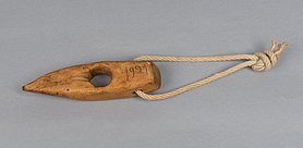 Das Seilholz sieht aus wie ein Keil. In der Mitte ist ein Loch. An dem Loch sind Spuren von Seilen. Auf der Seite steht die Jahreszahl 1921. In einem weiteren Loch ist eine Schlaufe. Mit ihr kann das Holz aufgehängt werden.