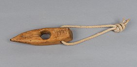 Das Seilholz sieht aus wie ein Keil. In der Mitte ist ein Loch. An dem Loch sind Spuren von Seilen. Auf der Seite steht die Jahreszahl 1921. In einem weiteren Loch ist eine Schlaufe. Mit ihr kann das Holz aufgehängt werden.