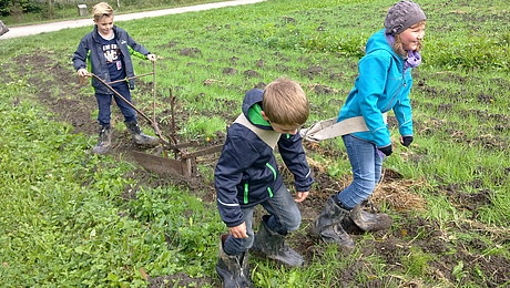 FFM_Kinder_beim_Pflügen Fotoaufnahme dreier Kinder beim Pflügen eines Feldes. Hinter dem alten Pflug steht ein Junge. Davor wurden mit Gurten ein weiterer Junge und ein Mädchen gespannt, die den Pflug durch die Erde ziehen. Im Hintergrund sind ein Weg und einige Bäume erkennbar.