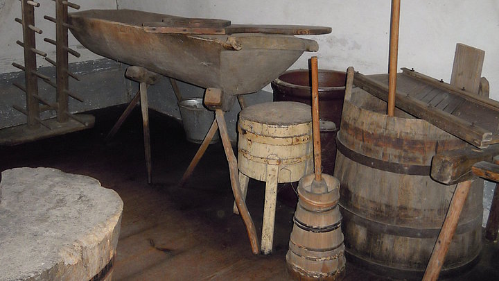 Fotoaufnahme der Vorratskammer im Bauernhaus aus Seubersdorf. Im Raum stehen verschiedene Geräte, die der Lebensmittelverarbeitung dienen. Gezeigt werden u.a. ein hölzernes Butterfass, ein Bottich für Kraut und ein Backtrog. An der Wand dahinter sind Malereien erkennbar. 