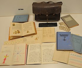 eine lederne Schultasche, davor zahlreiche Hefte und Schulbücher mit handschriftlichen Eintragungen.