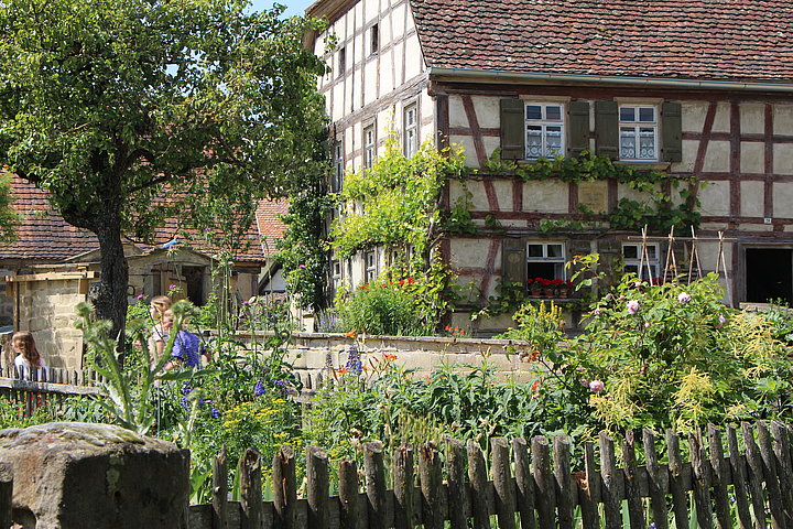 FFM_Gaerten Fotoaufnahme einer der Gärten des Museums. Im Vordergrund befindet sich der umzäunte Garten mit den blühenden Blumen im Inneren. Dahinter steht das Bauernhaus aus Herrnberchtheim, an der Wand des Fachwerks wächst Wein. Links daneben sind ein Baum und Besucher erkennbar.