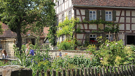 FFM_Gaerten Fotoaufnahme einer der Gärten des Museums. Im Vordergrund befindet sich der umzäunte Garten mit den blühenden Blumen im Inneren. Dahinter steht das Bauernhaus aus Herrnberchtheim, an der Wand des Fachwerks wächst Wein. Links daneben sind ein Baum und Besucher erkennbar.