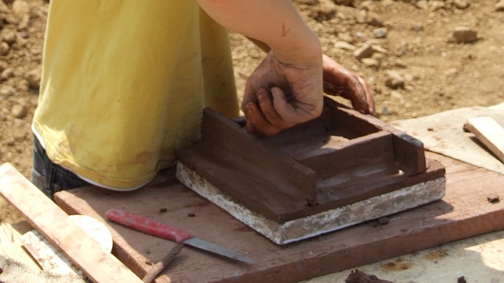 FFM_Ofenkachel Fotoaufnahme der Ofenkachelwerkstatt. Ein Junge mit T-Shirt und Hut steht an einem Holztisch. Auf einem Brett liegt eine Ofenkachel nach historischem Vorbild, die er gerade mit seinen Händen bearbeitet. Im Hintergrund befindet sich ein Dreckhaufen.