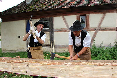 FFM_Handwerkertag Fotoaufnahme vom Handwerkertag. Gezeigt werden zwei Zimmermännern in Kluft, die einen aufgebockten Holzstamm mit Beilen bearbeiten. Im Hintergrund steht ein mit Flachziegeln gedecktes Fachwerk.