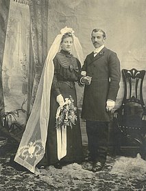 Ehepaar. Die Braut trägt ein schwarzes Kleid. Dazu trägt sie einen weißen Schleier. Der Bräutigam trägt einen schwarzen Anzug.