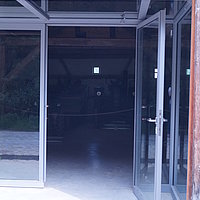 FFM_West_Weiltingen_Eingang Fotoaufnahme vom verglasten Eingangsbereich der Schafscheuer aus Weiltingen am aktuellen Standort. Im Gebäude sind der Sichtdachstuhl, sowie einige Maschinen erkennbar. Der Bereich der Exponate wird mit einem Seil abgegrenzt.