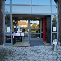 FFM_Eingangsbereich_Ausstellungsscheune Fotoaufnahme des Eingangsbereichs zur Ausstellungsscheune bzw. dem Stadel aus Betzmannsdorf. Das zweiflüglige Holztor ist nach außen geöffnet, dahinter liegt der verglaste Eingangsbereich mit geschlossener Tür. Auf dem grob gepflasterten Hof davor steht ein Aschenbecher.