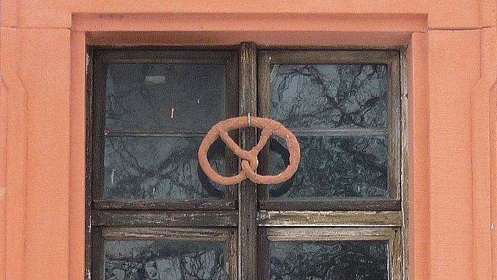 FFM_Backstube_Brezen_backen Fotoaufnahme des Fensters der Backstube im Amtshaus aus Obernbreit. Das Fenster ist in einem steinernen Rahmen eingebettet. Am hölzernen Fensterkreuz ist eine Brezel angebracht.