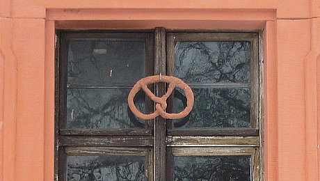 FFM_Backstube_Brezen_backen Fotoaufnahme des Fensters der Backstube im Amtshaus aus Obernbreit. Das Fenster ist in einem steinernen Rahmen eingebettet. Am hölzernen Fensterkreuz ist eine Brezel angebracht.