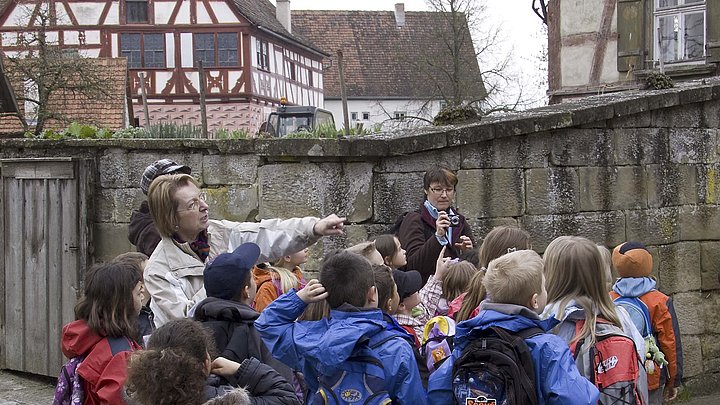 FFM_Frlm_Schlag Fotoaufnahme einer Gruppe Schulkinder, die vor dem Bauernhaus aus Herrnberchtheim steht. Hinter der Hofmauer ist neben kahlen Bäumen unter anderen das Amtshaus aus Obernbreit erkennbar. Die Führerin zeigt auf etwas außerhalb des Bildes.