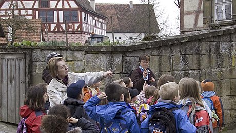 FFM_Frlm_Schlag Fotoaufnahme einer Gruppe Schulkinder, die vor dem Bauernhaus aus Herrnberchtheim steht. Hinter der Hofmauer ist neben kahlen Bäumen unter anderen das Amtshaus aus Obernbreit erkennbar. Die Führerin zeigt auf etwas außerhalb des Bildes.