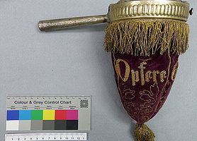 Der Beutel ist aus dunkelrotem Stoff. Auf ihm sind mit goldfarbenem Faden Blätter aufgestickt. Auch ist ein Schriftzug aufgestickt. Da steht "Opfere Gott Dank". Oben auf dem Beutel ist ein Ring mit einem Handgriff. Der Ring hält den Beutel offen.