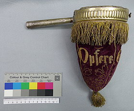Der Beutel ist aus dunkelrotem Stoff. Auf ihm sind mit goldfarbenem Faden Blätter aufgestickt. Auch ist ein Schriftzug aufgestickt. Da steht "Opfere Gott Dank". Oben auf dem Beutel ist ein Ring mit einem Handgriff. Der Ring hält den Beutel offen.