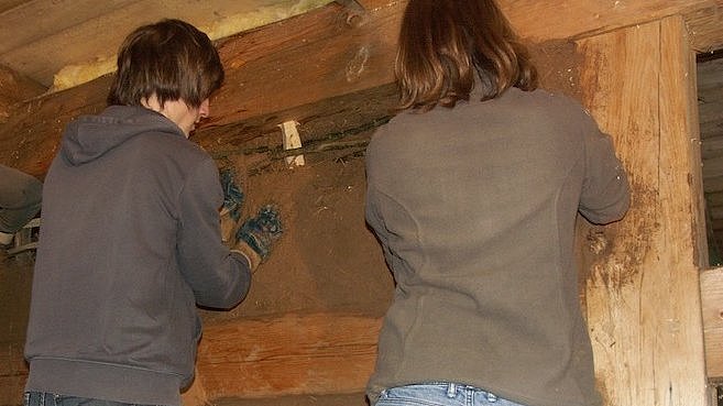 FFM_Aktionstage_Bauhandwerk Fotoaufnahme der Aktionstage „Bäuerliches und handwerkliches Arbeiten“. Zwei Jugendliche stehen vor einer Fachwerkwand und fachen diese mit einem Lehm-Strohgemisch aus. Der Großteil der Wand ist bereits ausgefacht. Beide Jungen tragen Jeans und Pullover.