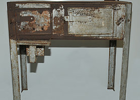 Fotoaufnahme des Schrottofens aus Ergersheim. Der metallene Ofen wurde aus Schrottteilen zusammengebaut. Er steht auf vier Beinen und hat zwei nebeneinanderliegende Klappen, die im Moment geschlossen sind. Hinten rechts ist ein kurzes Ofenrohr angebracht. 