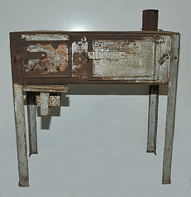 Fotoaufnahme des Schrottofens aus Ergersheim. Der metallene Ofen wurde aus Schrottteilen zusammengebaut. Er steht auf vier Beinen und hat zwei nebeneinanderliegende Klappen, die im Moment geschlossen sind. Hinten rechts ist ein kurzes Ofenrohr angebracht. 