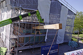 7.5.18 Das Einbringen der Deckenbauteile, hier einer der Unterzüge für die Badstube, erfolgt mit Hilfe eines Teleskopladers seitlich durch das Gerüst des Schutzdaches.