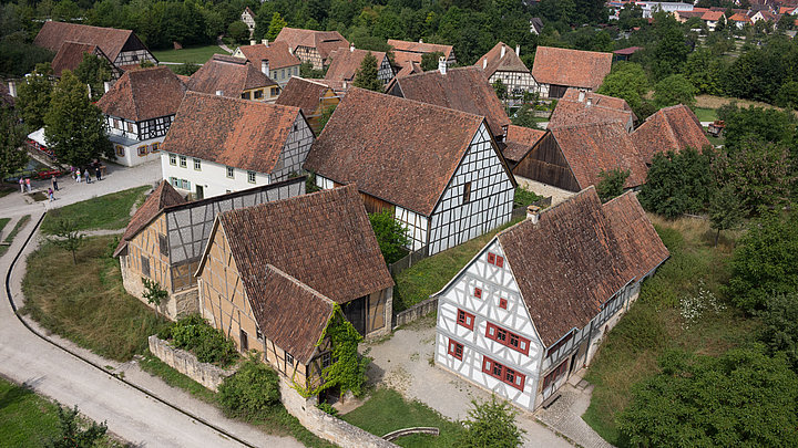 FFM_Mainfranken_Frankenhöhe_Luftaufnahme Fotoaufnahme des Luftbildes der Baugruppe West. Die Gebäude, zum Großteil Fachwerke, stehen nah beieinander und sind von Gärten, Wiesen, Bäumen und Wegen umgeben. Im Hintergrund sind die weitere Landschaft des Museums sowie Bad Windsheims erkennbar.