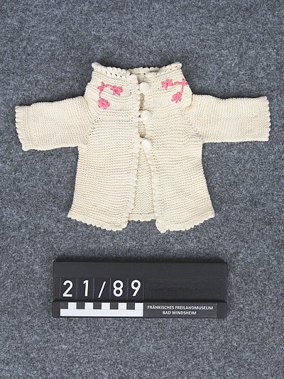 Die kleine Jacke ist gestrickt. Sie ist beige. Auf dem Kragen sind rosafarbene Blumen.