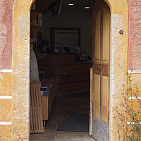 FFM_Eingang_Gelände Fotoaufnahme des Eingangs zum Gelände. Im Raum dahinter ist die Museumskasse erkennbar, sie befindet sich im Korbhaus aus Knittelsbach. Über der geöffneten Holztür steht „JAL 1812“.