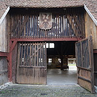 FFM_Vorderer_Eingang_Alter_Bauhof Fotoaufnahm des vorderen Eingangs zum Alten Bauhof aus Bad Windsheim. Die rechte Seite des zweiflügligen Holztors ist geöffnet. Darüber hängt ein Wappen mit einem Adler und einem „W“ in der Mitte. Im Inneren sind einige Objekte erkennbar.