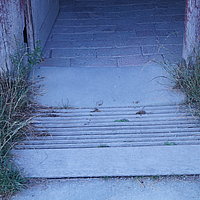 FFM_West_Schaeferei_Rampe_4 Fotoaufnahme der Schwelle am linken Eingang der Schäferei aus Hambühl am aktuellen Standort. Vor der Schwelle liegt eine Rampe aus Holz, links und rechts davon wächst Gras. Der Boden im Inneren ist gepflastert.
