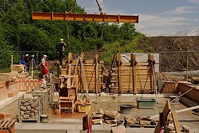 Mauerwerksteile sind an Position Fotoaufnahme zweier Mauerwerksteile mit Verschalung an ihrer endgültigen Position im Erdgeschoss. Um das linke Bauteil, welches noch am Kran befestigt ist, stehen mehrere Arbeiter mit Schutzhelmen. Hinter dem Bauzaun sind Bäume und ein Erdhügel erkennbar.