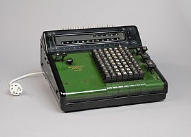 Die Rechenmaschine ist grün.Sie hat 64 Tasten mit Zahlen. Man kann mit ihr rechnen. Die Rechenergebnisse sieht man auf zwei Leisten. Sie hat weitere Tasten zur Bedienung. Sie funktioniert mit Strom. 