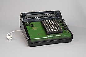Die Rechenmaschine ist grün.Sie hat 64 Tasten mit Zahlen. Man kann mit ihr rechnen. Die Rechenergebnisse sieht man auf zwei Leisten. Sie hat weitere Tasten zur Bedienung. Sie funktioniert mit Strom. 