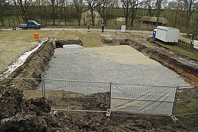 FFM_Bautagebuch_Wendelstein_24_3_17 Fotoaufnahme der Baugrube mit der eingebauten Schotterlage, die aus Schrobben und Grobschotter besteht. Die Baustelle ist von einem Bauzaun umgeben. Rechts steht ein Bauwagen und ein Dixi. Im Hintergrund sind eine Reihe kahler Bäume sowie einige Gebäude erkennbar.