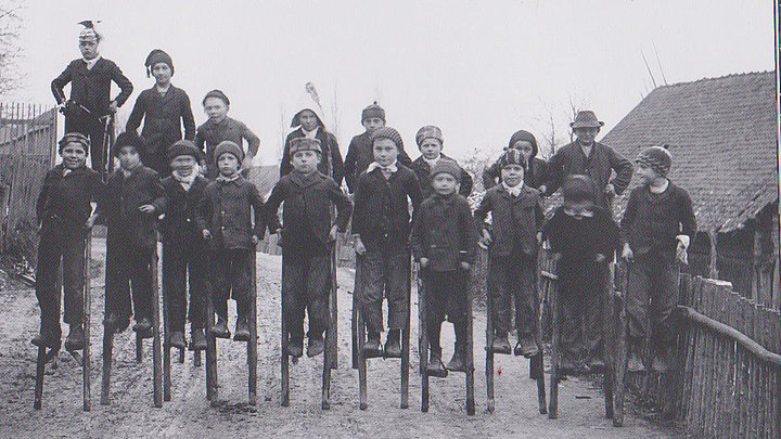 FFM_Stelzen Historische schwarz-weiße Fotoaufnahme einer 20-köpfigen Kindergruppe auf hölzernen Stelzen. Sie stehen in zwei Reihen hintereinander auf einem Feldweg in einem Dorf. Alle Kinder tragen Jacken und Mützen. Links im Hintergrund sind kahle Bäume erkennbar, rechts ein Haus.