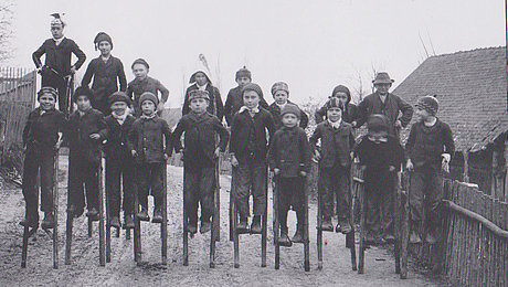 FFM_Stelzen Historische schwarz-weiße Fotoaufnahme einer 20-köpfigen Kindergruppe auf hölzernen Stelzen. Sie stehen in zwei Reihen hintereinander auf einem Feldweg in einem Dorf. Alle Kinder tragen Jacken und Mützen. Links im Hintergrund sind kahle Bäume erkennbar, rechts ein Haus.