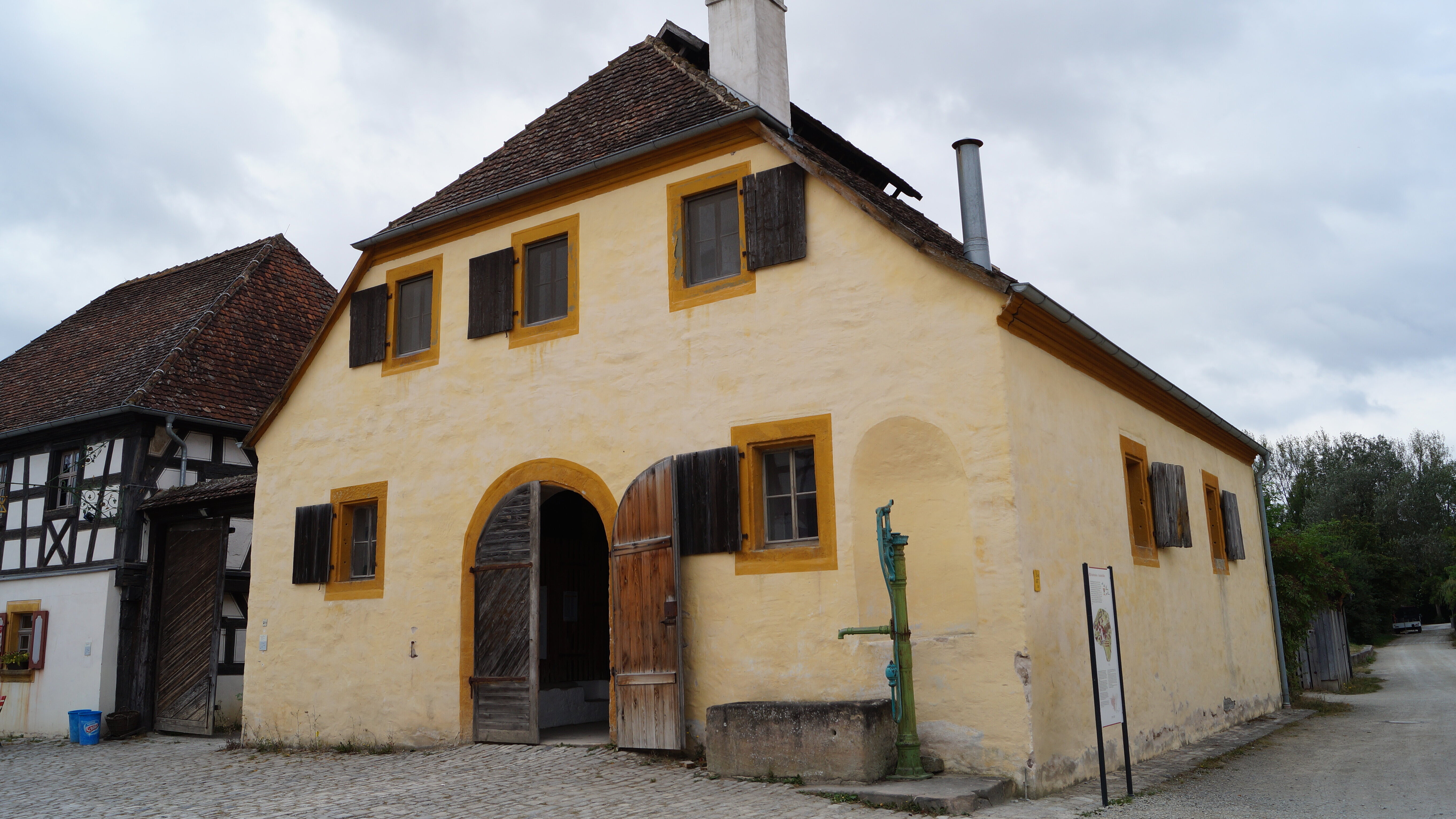 FFM_Kommunbrauhaus_aus_Schlüsselfeld Fotoaufnahme des Kommunbrauhauses aus Schlüsselfeld am aktuellen Standort. Rechts vor dem verputzen Bruchsteinbau steht ein Pumpbrunnen mit Steintrog. Links neben dem Gebäude ist die Wirtschaft am Brauhaus erkennbar. Der Himmel ist wolkenverhangen.