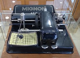 Das Foto zeigt eine schwarze Schreibmaschine. Sie besitzt einen Zeiger, den man zu den Buchstaben und Zeichen führen kann.