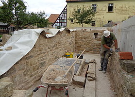 niedrig aufgemauerte Sansteinmauern, darin arbeitet ein Maurer. Schubkarre und Werkzeug umgibt ihn.