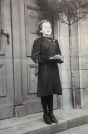Auf dem Bild ist ein Mädchen. Es trägt ein Kleid und ein Gesangbuch. Es hat schwarze Schuhe an. Es steht vor einer Kirchentür.