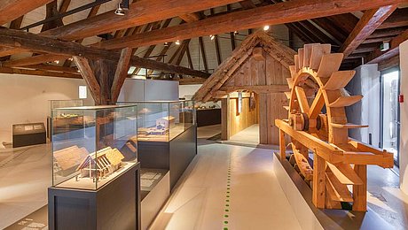 FFM_Ausstellung_Archaeologische_Staatssammlung Fotoaufnahme des Innenraums vom Archäologiemuseum in der Schafscheune aus Virnsberg. Ein Bodenleitsystem führt durch die Ausstellung. Links vom Weg stehen Vitrinen mit Modellen. Rechts ein hölzerner Wasserrad. Dahinter ist eine kleine Hütte aus Holz aufgebaut.