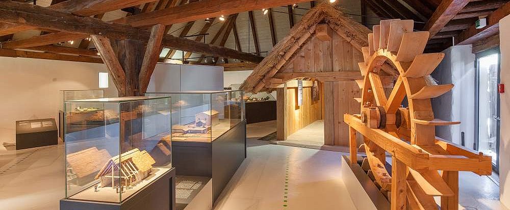FFM_Ausstellung_Archaeologische_Staatssammlung Fotoaufnahme des Innenraums vom Archäologiemuseum in der Schafscheune aus Virnsberg. Ein Bodenleitsystem führt durch die Ausstellung. Links vom Weg stehen Vitrinen mit Modellen. Rechts ein hölzerner Wasserrad. Dahinter ist eine kleine Hütte aus Holz aufgebaut.