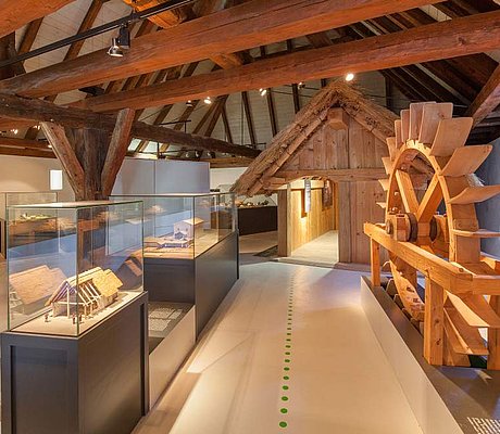 FFM_Ausstellung_Archaeologische_Staatssammlung Fotoaufnahme des Innenraums vom Archäologiemuseum in der Schafscheune aus Virnsberg. Ein Bodenleitsystem führt durch die Ausstellung. Links vom Weg stehen Vitrinen mit Modellen. Rechts ein hölzerner Wasserrad. Dahinter ist eine kleine Hütte aus Holz aufgebaut.