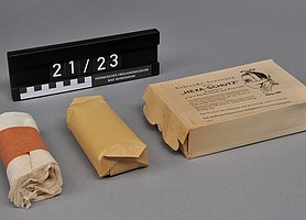 Die Maske ist eingewickelt. Die Schachtel ist braun. Auf der Schachtel ist ein Text. Der Text zeigt, wie man die Maske benutzt.