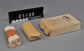 Die Maske ist eingewickelt. Die Schachtel ist braun. Auf der Schachtel ist ein Text. Der Text zeigt, wie man die Maske benutzt.