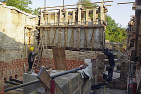 Die Innenwand trennt Badstube und Vorbad Fotoaufnahme des Einsetzens der Innenwand, die Badstube und Vorbad trennt. Auf das Mauerwerk aus Bruchstein und Ziegel wird mit einem Schwerlastkran das verschalte Wandstück gesetzt. Arbeiter am Boden helfen bei der Positionierung.