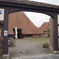 FFM_Eingang_Alter_Bauhof Fotoaufnahme des Eingangs in den Hof des Alten Bauhofs. Das zweiflügelige Holztor ist nach innen geöffnet. Im Hintergrund ist der Bauhofstadel aus Bad Windsheim erkennbar, davor stehen mehrere Tische und Stühle und ein zusammengeklappter Sonnenschirm. Der Boden ist gepflastert.