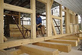 Fotoaufnahme der aufgestellten östlichen Fachwerktraufwand. Die Hohlräume zwischen den Holzbalken sind noch nicht ausgefüllt. In der Türöffnung steht ein Arbeiter, er hält einen Spanngurt in der Hand, der um die Türschwelle gewickelt ist. Im Hintergrund ist das Gerüst erkennbar. 