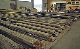 19.9.2017 Fotoaufnahme der Abbundhalle der Firma Lederer. In dieser wurde die bauzeitliche Balkenlage des Erdgeschosses sortiert und der Reihe nach auf dem Boden ausgelegt. Einige Holzbalken sind beschädigt. Das Tor der Halle ist geöffnet, an den Wänden befinden sich Regale und Baumaterialien.