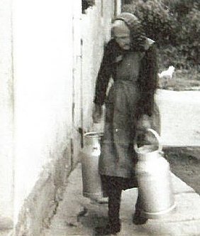 Eine hochbetagte Frau trägt in beiden Händen je eine Milchkanne, bewegt sich scheinbar vom Hof in den Stall?