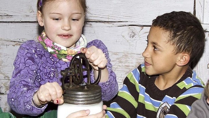 Fotoaufnahme der Führung „Von der Kuh zum Butterbrot“.  Um einen Tisch mit einem Butterglas sitzen Kinder. Das Glas wird von zwei Jungen festgehalten, von dem linken Jungen sieht man aber nur die Arme. Das Mädchen in der Mitte dreht an der Kurbel. 