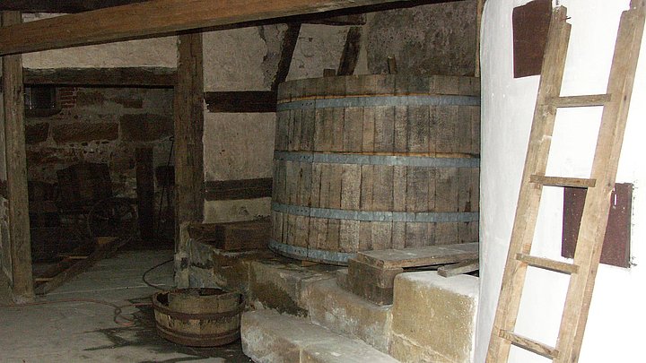 FFM_Kraisdorf_Brauerei Fotoaufnahme des Inneren des Hofbrauhauses aus Kraisdorf. Im vorderen Bereich befindet sich ein hoher Kessel mit Steinwand, an dem eine Holzleiter lehnt. Dahinter stehen ein hölzerner Sudkessel und ein Läuterbottich. Die Tür an der hintern Wand führt in den angrenzenden Raum.